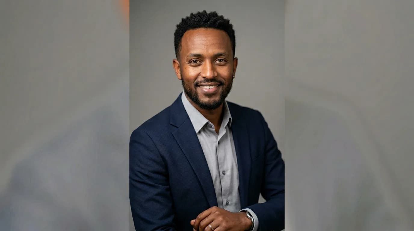 A man in a navy suit, smiling, photographed against a grey background